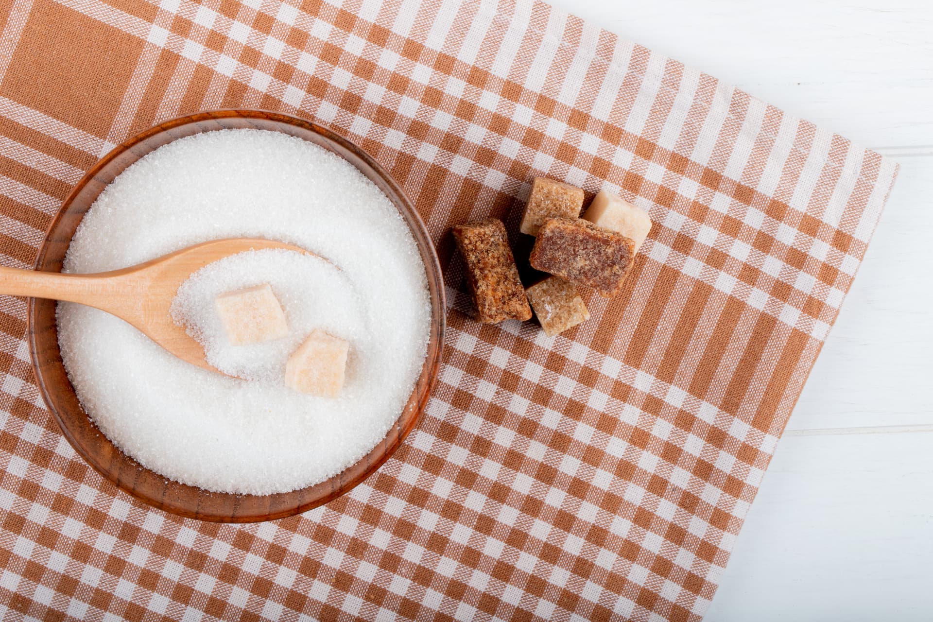 Bol en bois de sucre blanc avec une cuillère en bois et des morceaux de sucre, posé sur une nappe à carreaux beige et blanc