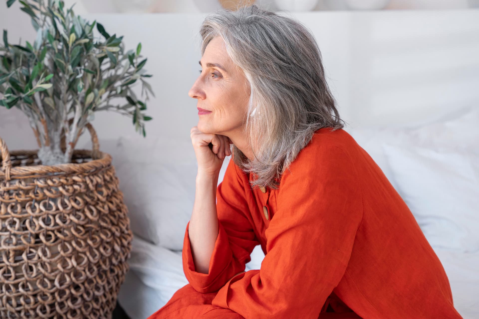 Femme d'une soixantaine d'années, cheveux gris, chemisier terracotta, posant en réflexion près d'une plante dans un intérieur clair